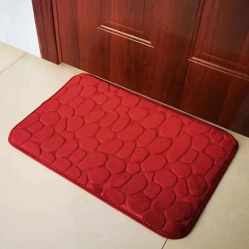 A wine red colored Memory Foam Bath Mat on the floor outside of a wooden door.