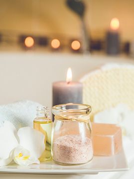 a white square plate holding a lit candle, a bath soap bar, a bottle of serum and a jar of bath salts